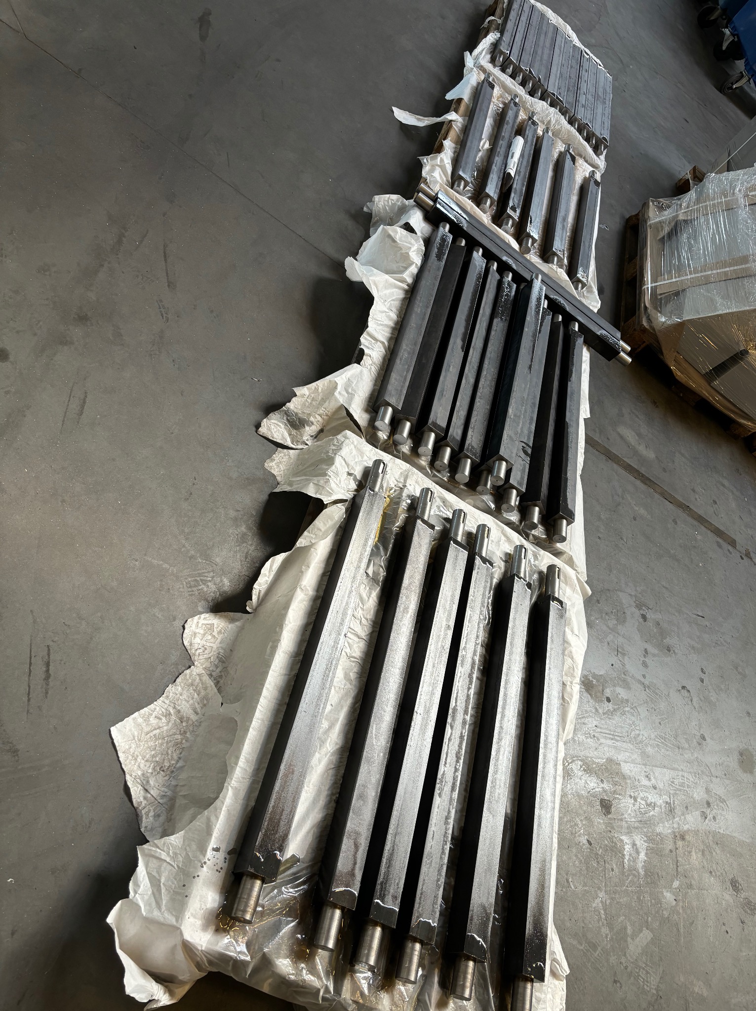 Czarne, metalowe pręty o przekroju kwadratowym z okrągłymi końcówkami, ułożone równolegle na białym papierze pakowym na szarej betonowej posadzce, prawdopodobnie po obróbce.