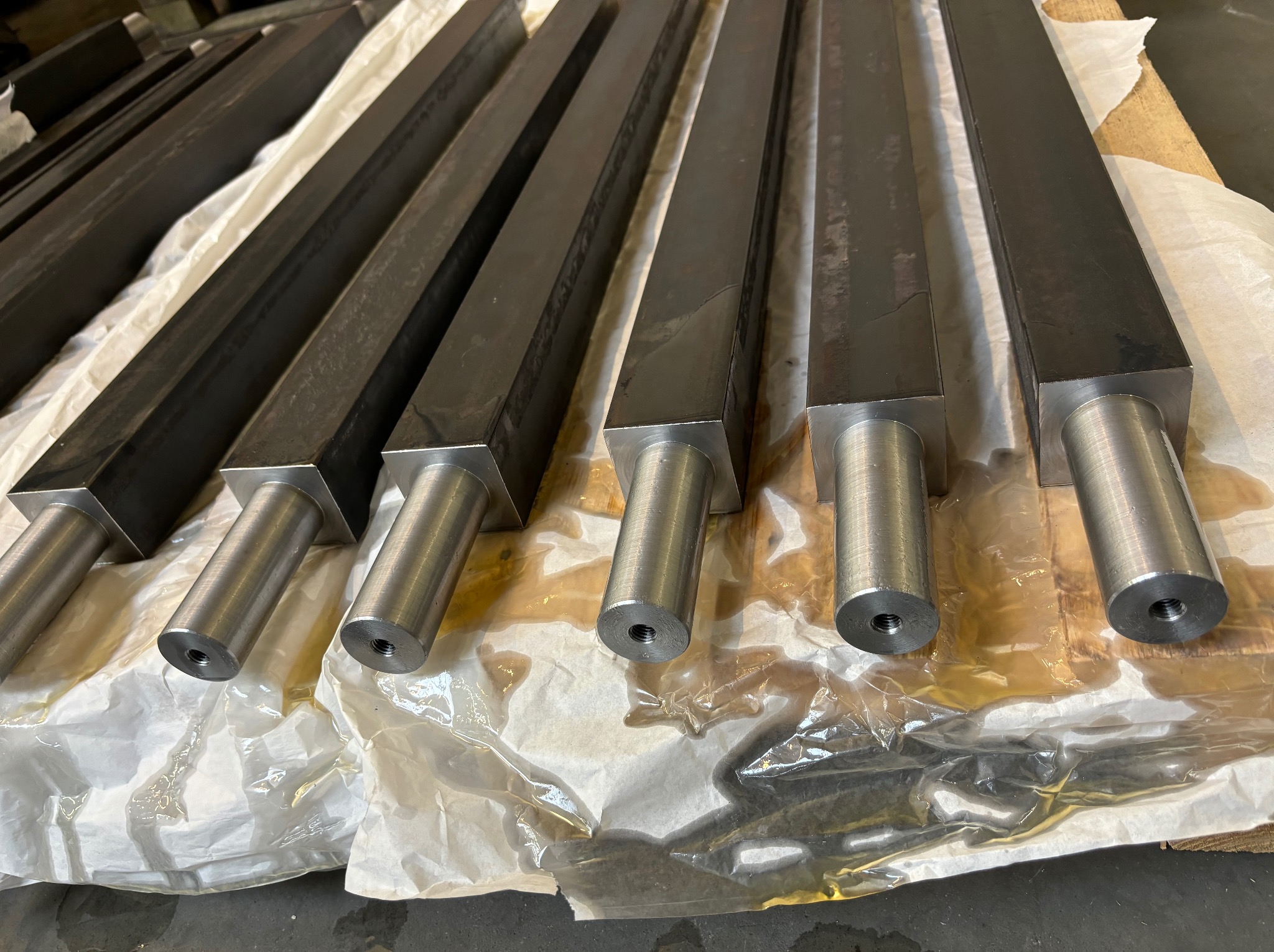 Seryjnie wykonane, metalowe elementy z czworokątną podstawą i cylindrycznym trzpieniem z gwintem wewnętrznym, ułożone na folii ochronnej z widocznymi śladami oleju.