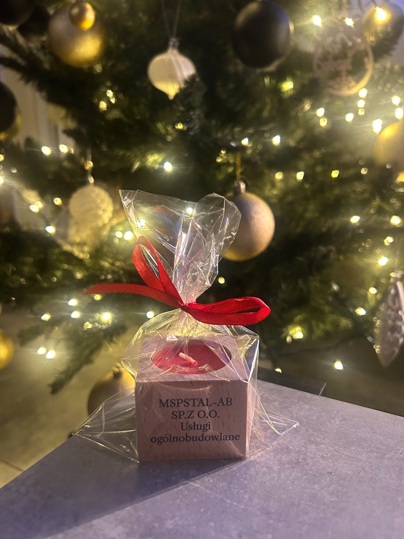 Świąteczna świeczka zapachowa w drewnianym pudełku z logo firmy MSPSTAL-AB SP.Z O.O. ogólnobudowlane, zapakowana w folię z czerwoną wstążką, na tle choinki z bombkami i światełkami.