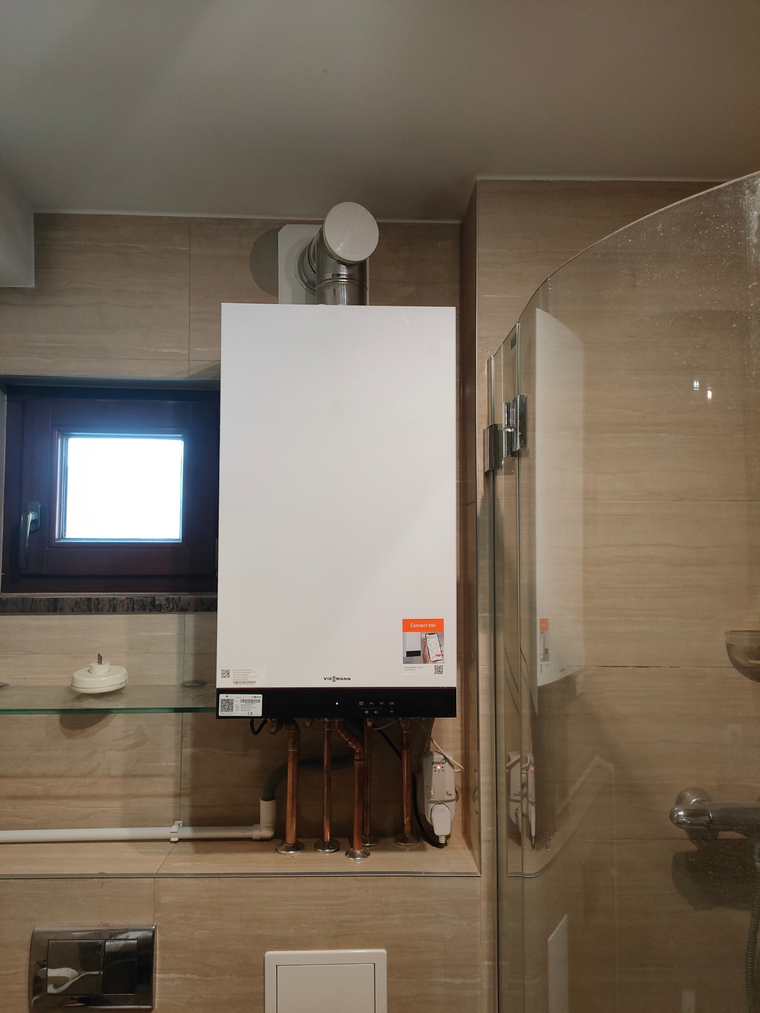 Nowoczesny, biały piec gazowy Viessmann zamontowany w łazience z beżowymi płytkami, widoczne miedziane rury instalacji, okno w tle i fragment kabiny prysznicowej.