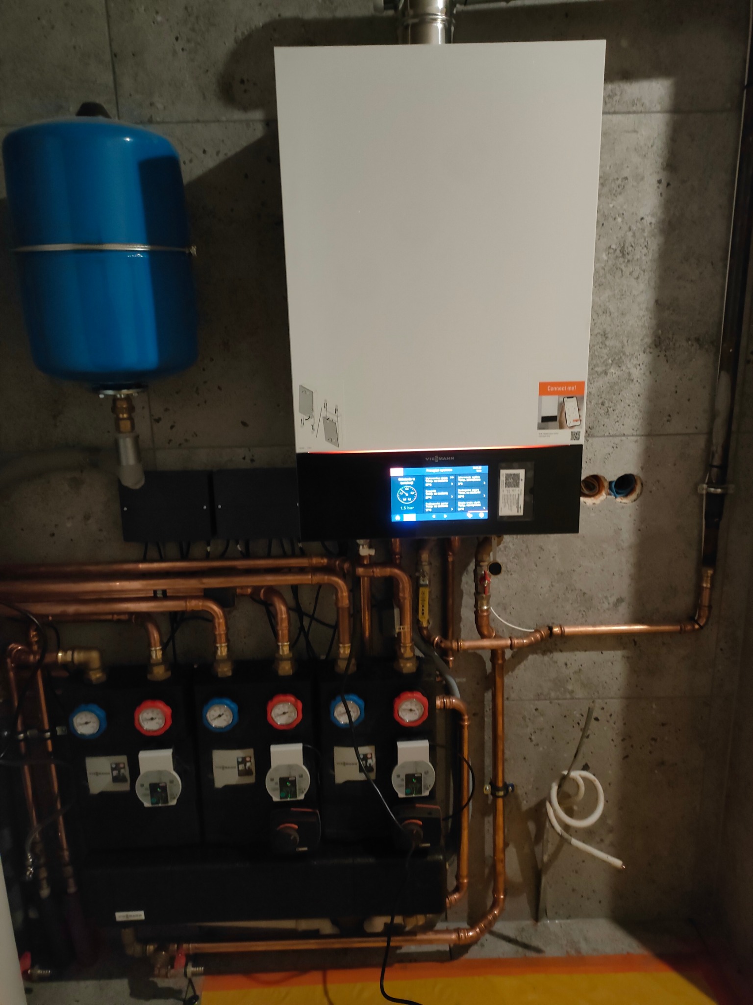 Nowoczesny piec gazowy Viessmann z elektronicznym wyświetlaczem, podłączony do instalacji z miedzianych rur i zbiornika wyrównawczego, zamontowany na szarej, betonowej ścianie.