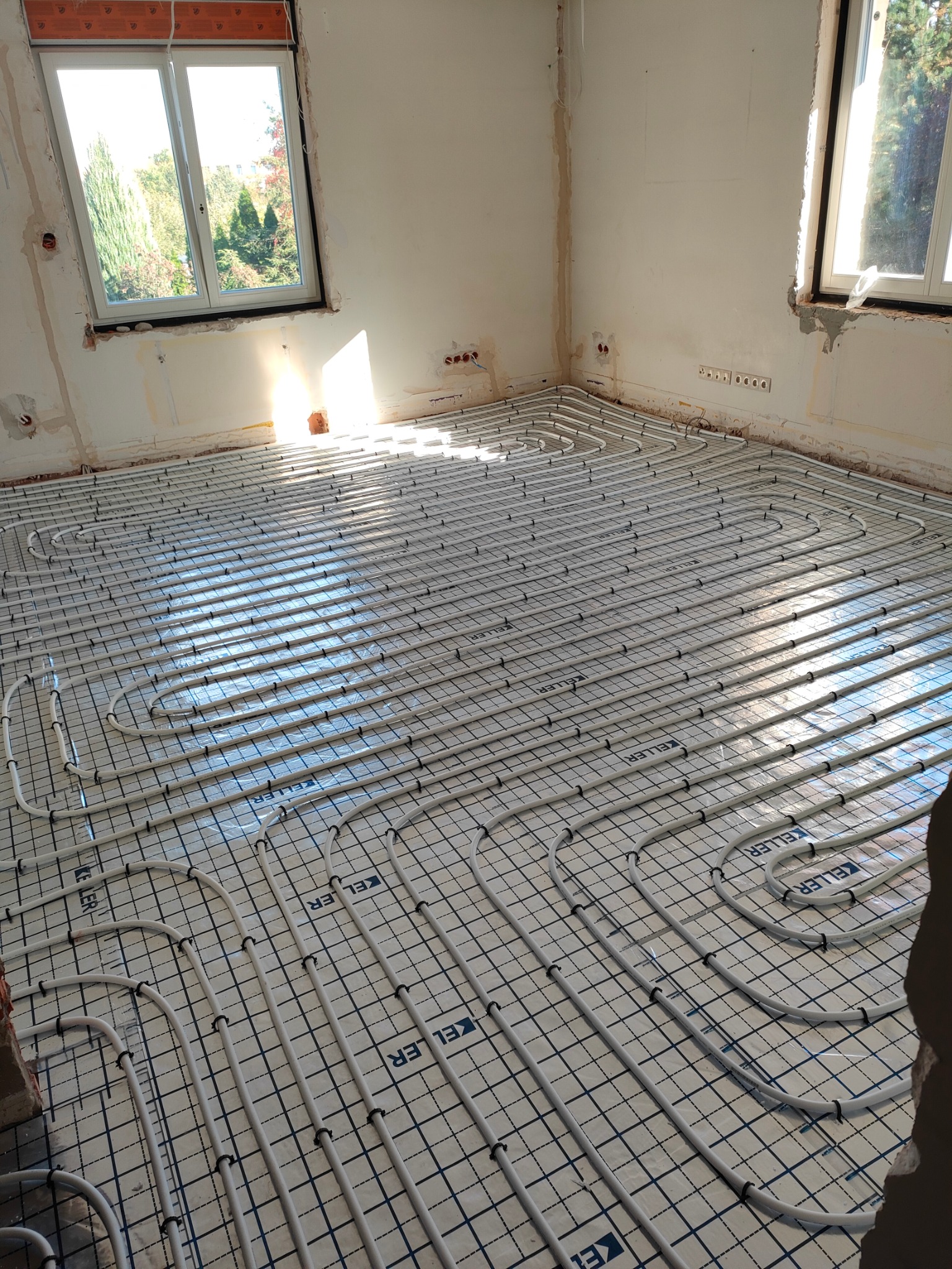 Widok na instalację ogrzewania podłogowego w trakcie montażu, rury ułożone na folii z nadrukiem siatki, surowy stan ścian, okno z widokiem na drzewa w tle.