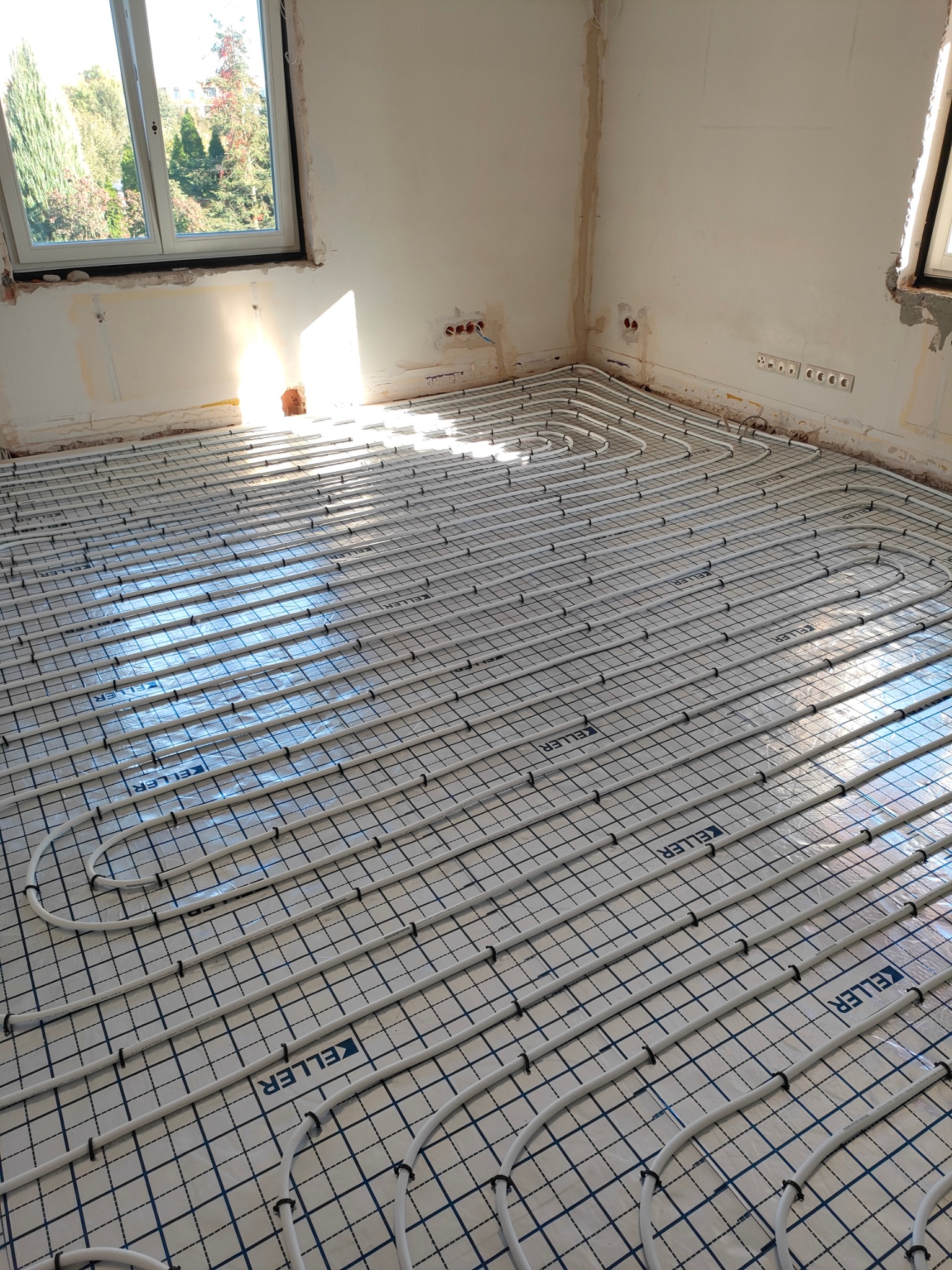 Białe rurki ogrzewania podłogowego ułożone na folii z nadrukiem kratki i logo 'KELLER', w surowym pomieszczeniu z oknem i widocznymi instalacjami elektrycznymi w ścianie.