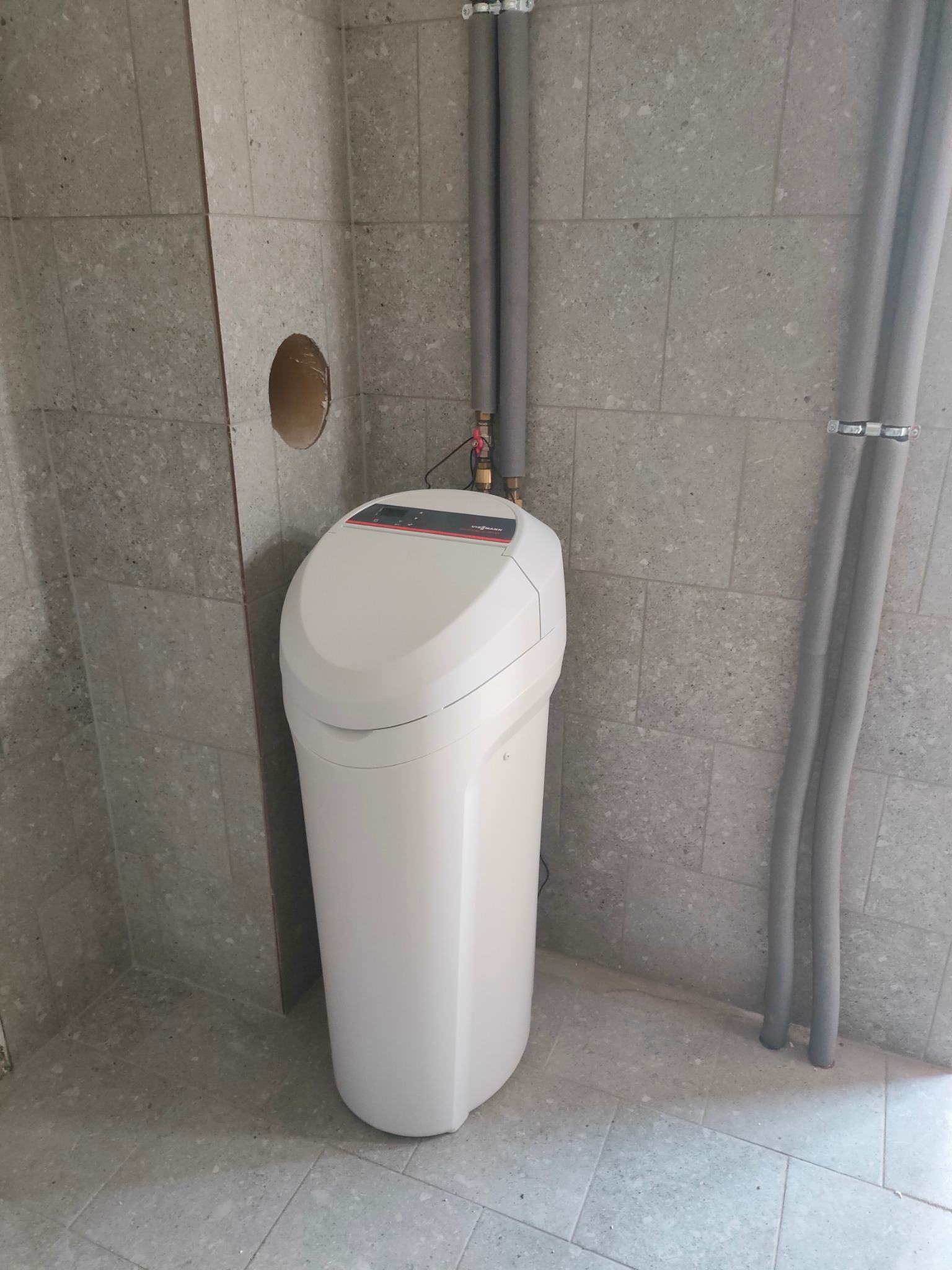 Biały zmiękczacz wody Viessmann w szarej kotłowni z rurami grzewczymi w izolacji, widoczny otwór w ścianie.