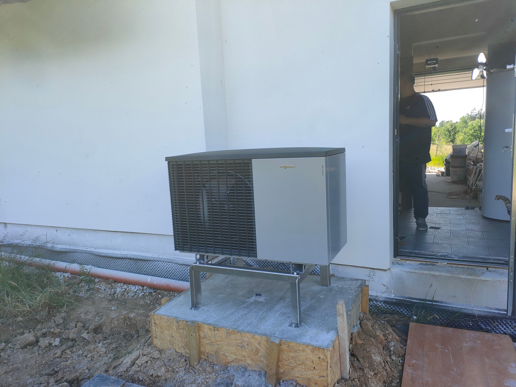 Zewnętrzna jednostka pompy ciepła Viessmann zamontowana na betonowym fundamencie z drewnianą szalunką, widoczna instalacja przyścienna i otwarte drzwi do budynku.