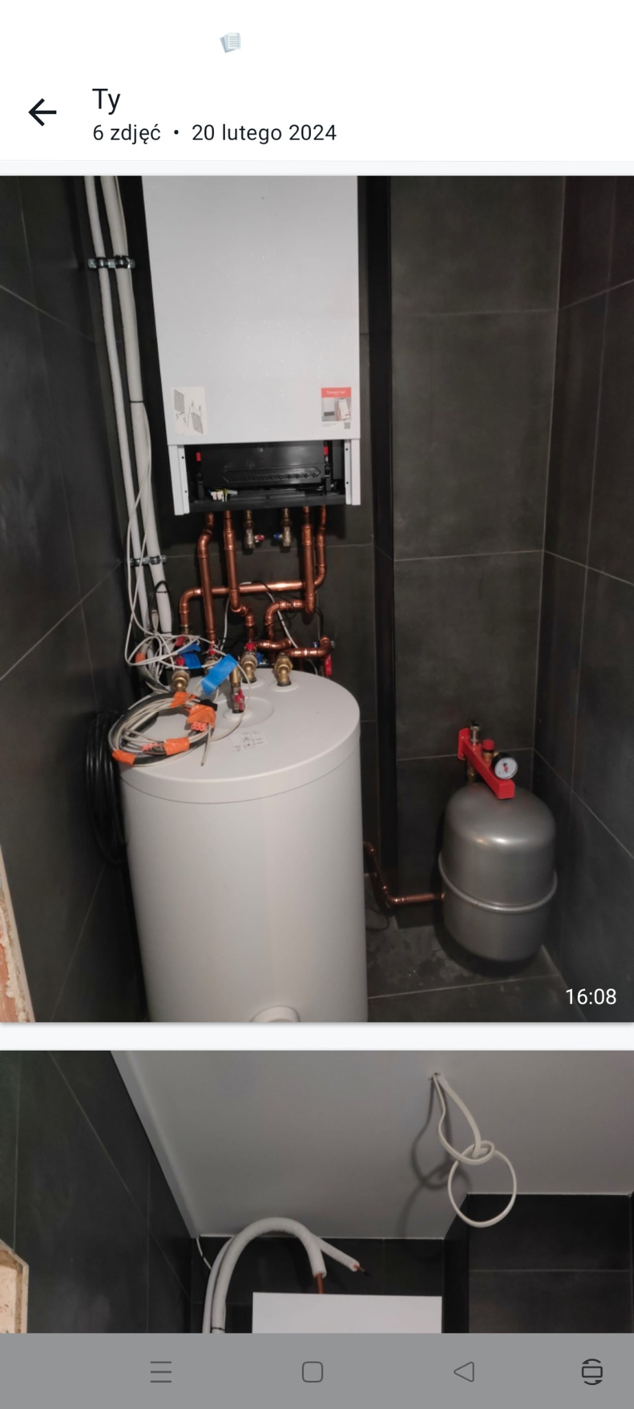 Biały piec gazowy kondensacyjny z podłączonym zasobnikiem ciepłej wody i naczyniem przeponowym w nowoczesnej łazience z ciemnymi płytkami, widoczne przewody i rury miedziane.