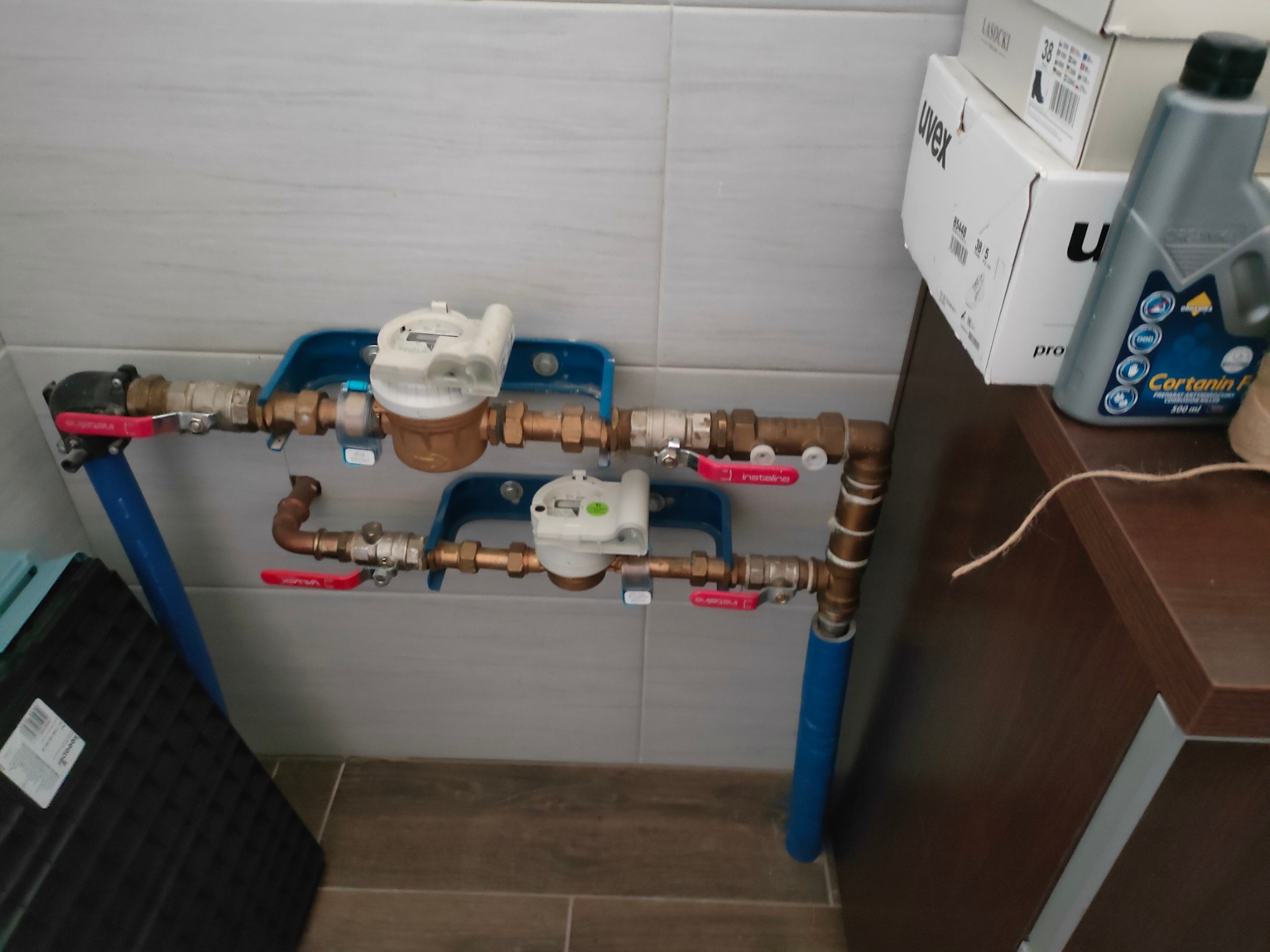 Dwa liczniki wody zamontowane na ścianie z szarych płytek, z widocznymi rurami miedzianymi, zaworami z czerwonymi rączkami i niebieskimi wspornikami. W tle widoczne pudełka i butelka płynu.