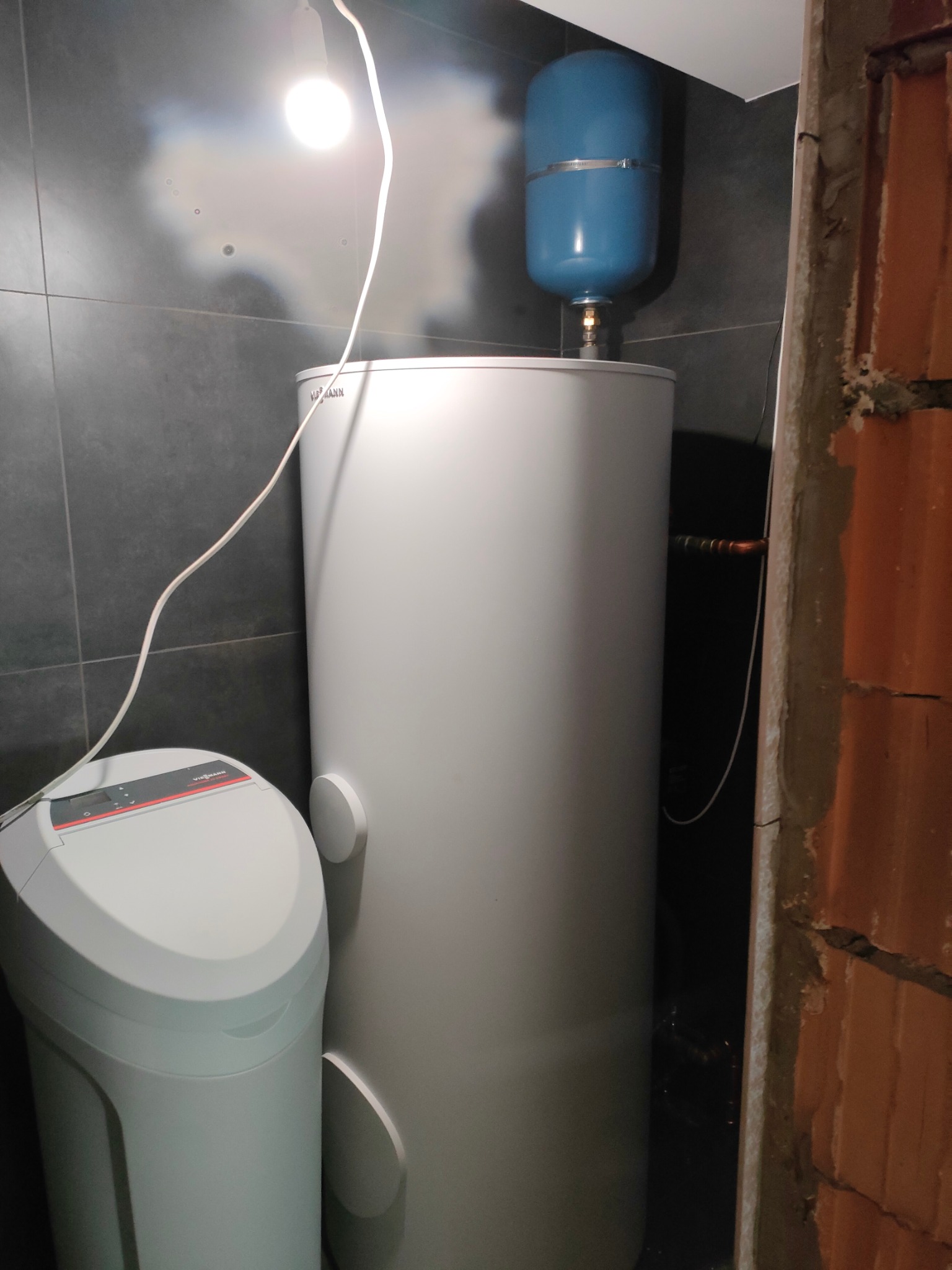 Biały zbiornik akumulacyjny Viessmann z niebieskim naczyniem wzbiorczym, zamontowany w kotłowni z szarymi płytkami i fragmentem surowej ściany z cegieł.