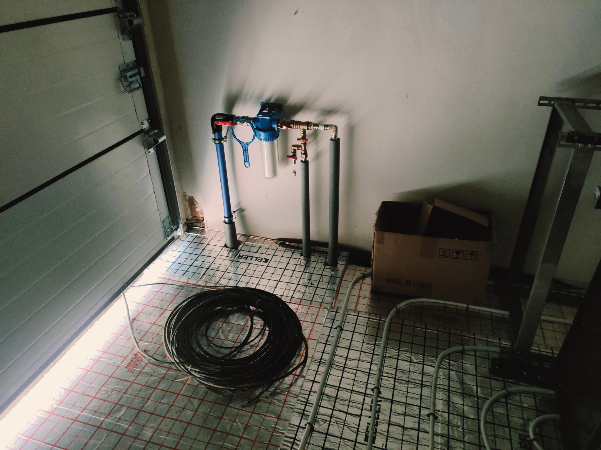 Filtr do wody zamontowany na rurach nad ogrzewaniem podłogowym w garażu z roletą segmentową.