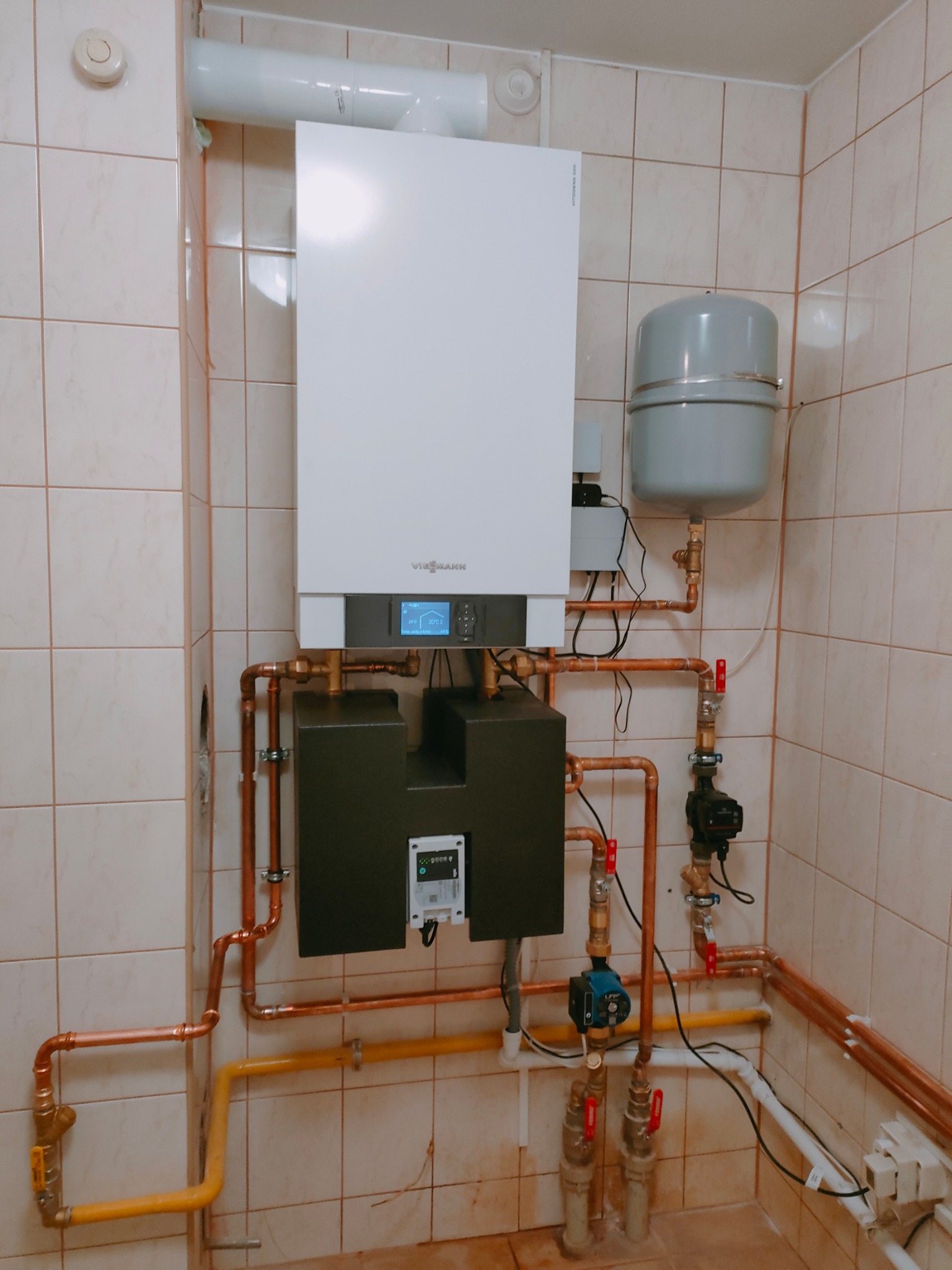 Biały piec gazowy marki Viessmann zamontowany na kafelkowanej ścianie z widocznymi miedzianymi rurami, zbiornikiem wyrównawczym i pompą obiegową.