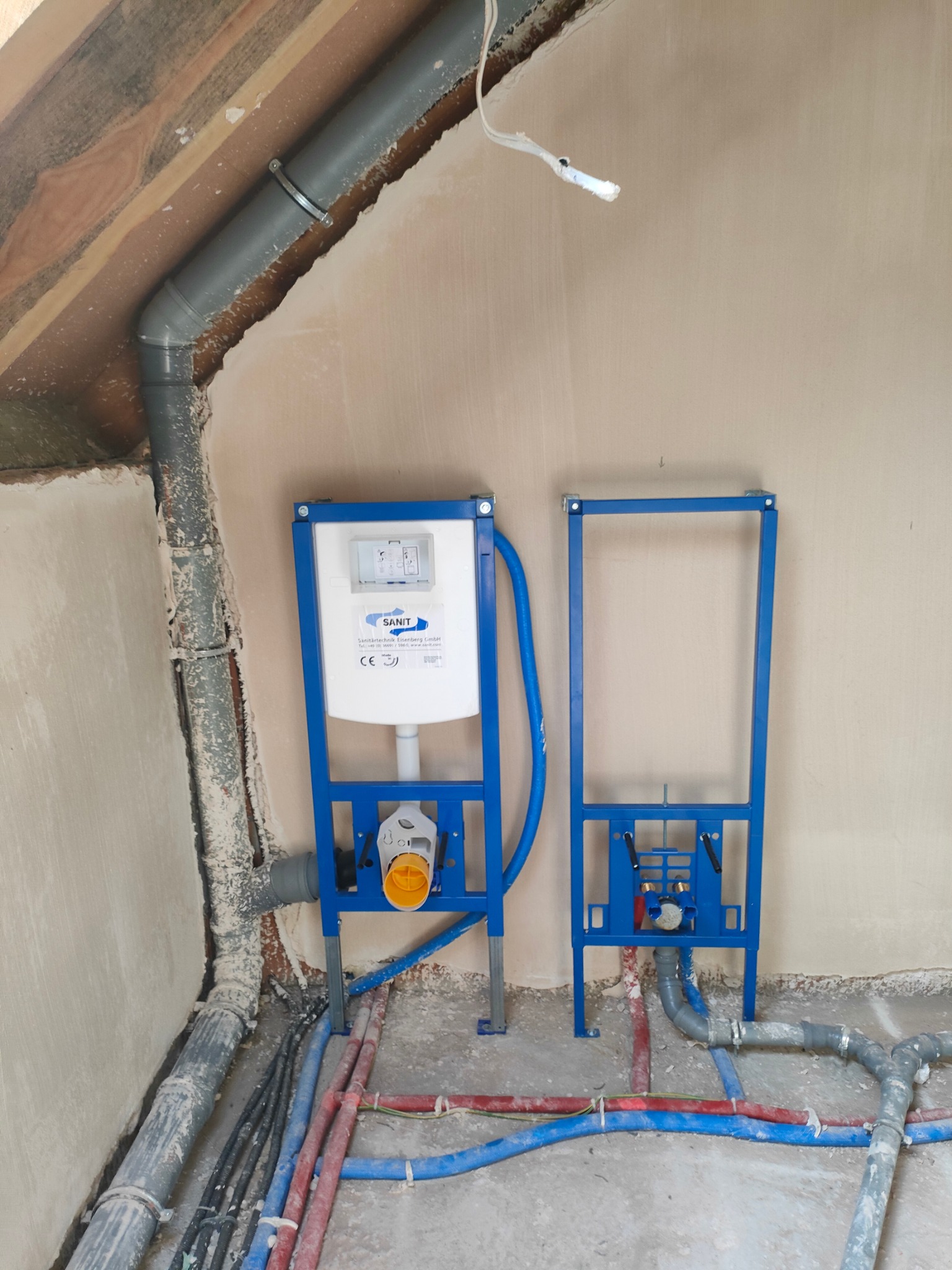 Stelaż podtynkowy WC marki Sanit zamontowany na tle surowej ściany i podłogi z widocznymi rurami instalacyjnymi. Widoczny fragment instalacji kanalizacyjnej pod sufitem.