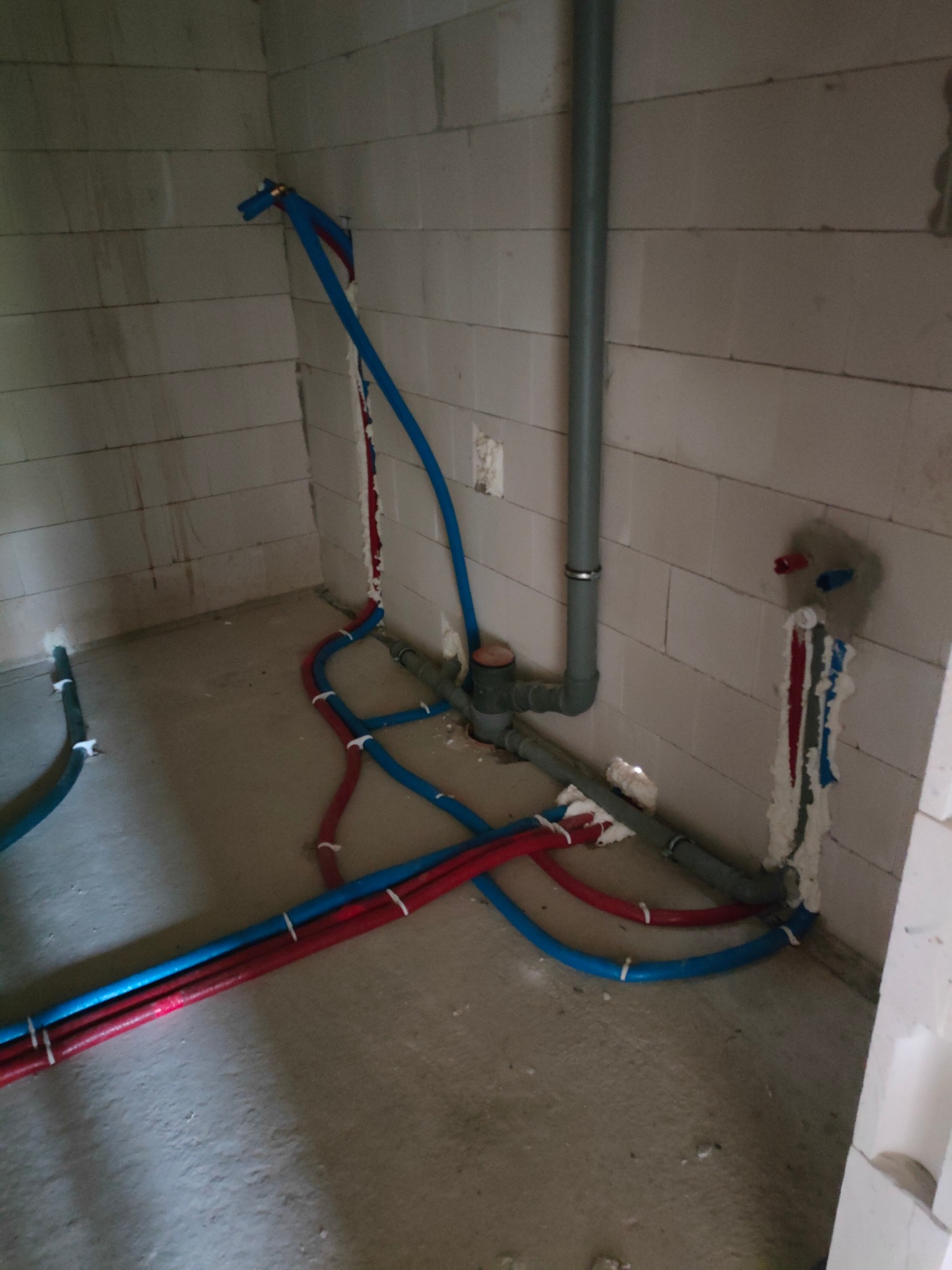 Surowy stan instalacji wodnej i kanalizacyjnej w budynku z bloczków, widoczne rury PEX w kolorze czerwonym i niebieskim oraz pion kanalizacyjny, uszczelnione pianką montażową w miejscach przejść...