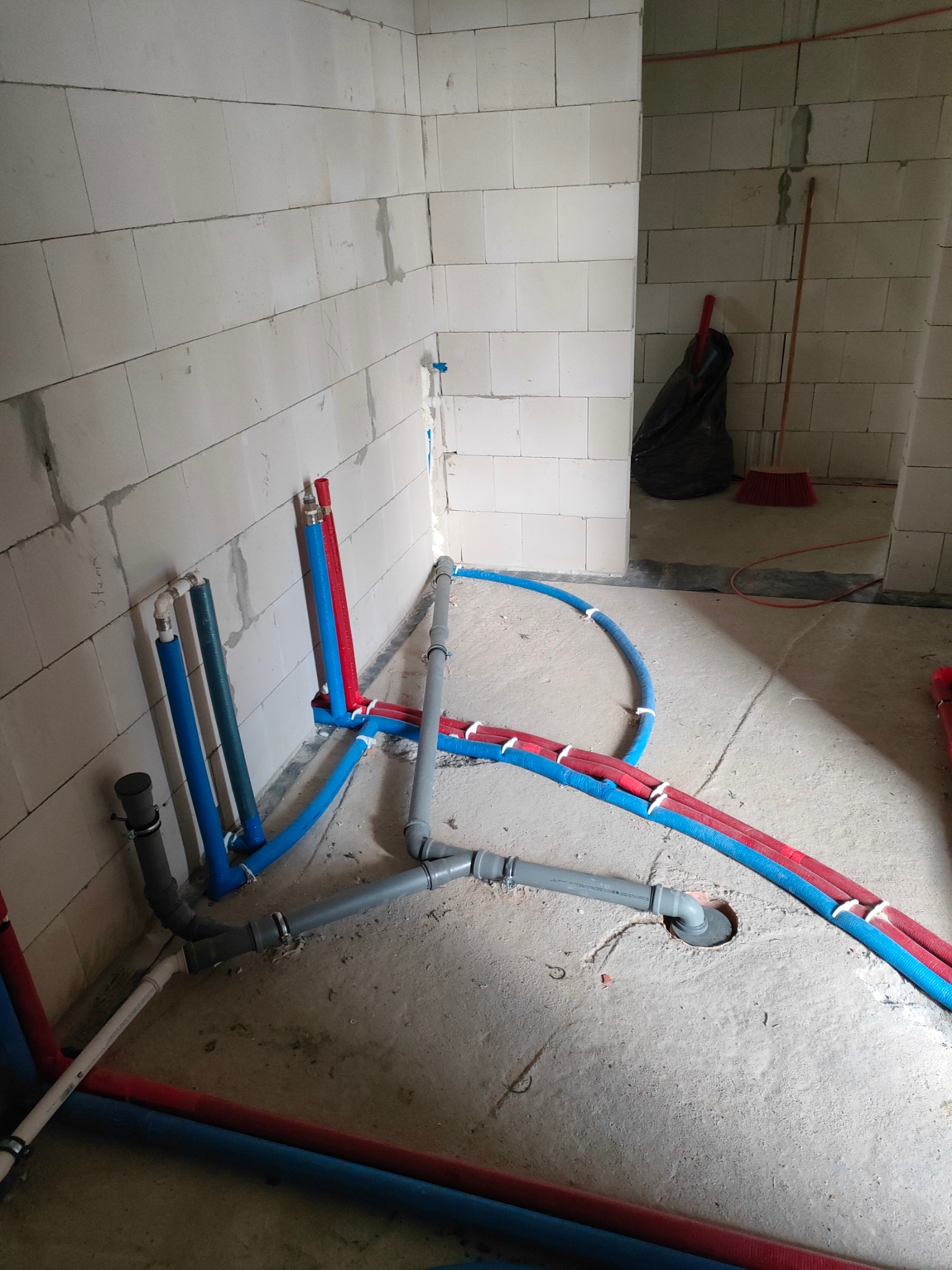 Wnętrze w trakcie budowy, widoczne rury instalacji wodnej i grzewczej w kolorach niebieskim i czerwonym, prowadzone po surowym betonowym podłożu i wzdłuż ściany z białych bloczków.