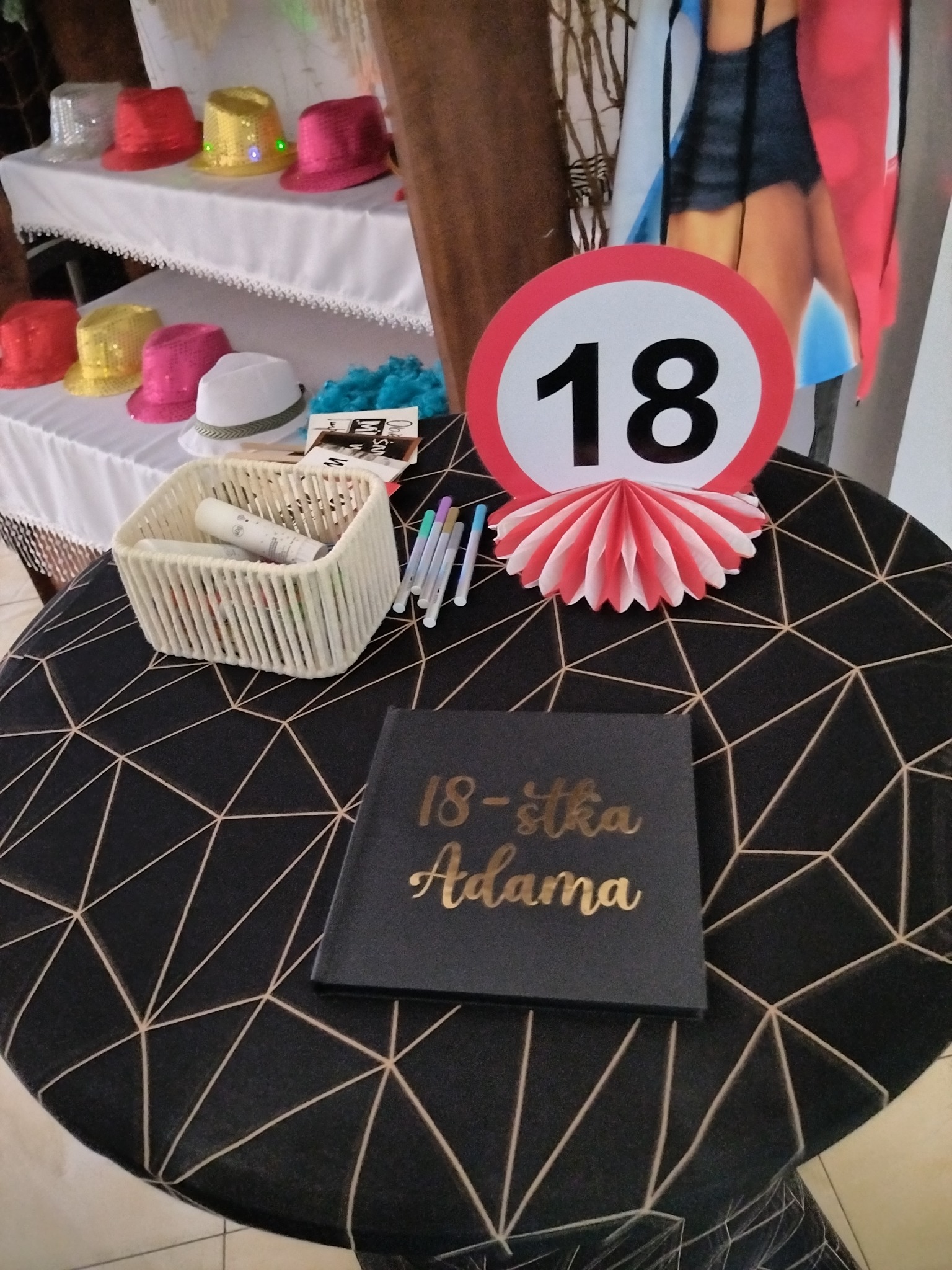 Dekoracje na 18 urodziny: księga gości z napisem '18-stka Adama', znak '18', koszyk z rekwizytami i kolorowe kapelusze na stole z geometrycznym wzorem.