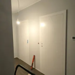 Dwa białe, minimalistyczne skrzydła drzwiowe zamontowane w szarej ścianie, poziomica oparta o framugę, widok z góry na rozłożony karton ochronny na podłodze i fragment drabiny malarskiej