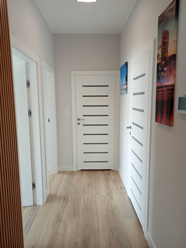 Korytarz z trzema białymi drzwiami o minimalistycznym designie, wykończony panelami podłogowymi w odcieniach beżu i szarości, z obrazami na ścianach.