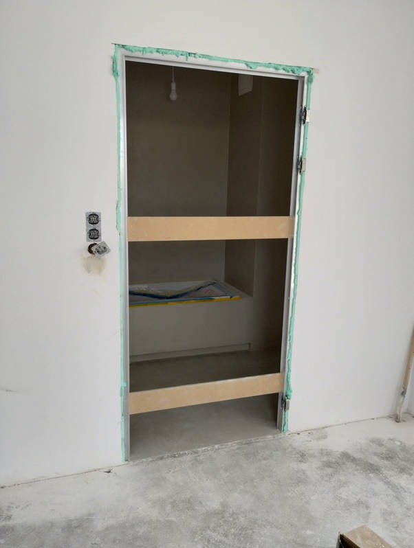 Ościeżnica drzwiowa zamontowana w surowym stanie, zabezpieczona drewnianymi rozpórkami, z widoczną pianką montażową i wystającymi przewodami elektrycznymi.