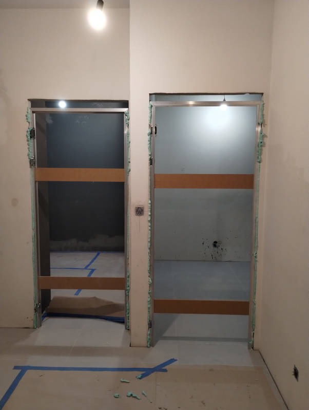 Dwa otwory drzwiowe z tymczasowymi zabezpieczeniami, pianką montażową i drewnianymi poprzeczkami, podłoga zabezpieczona niebieską taśmą.