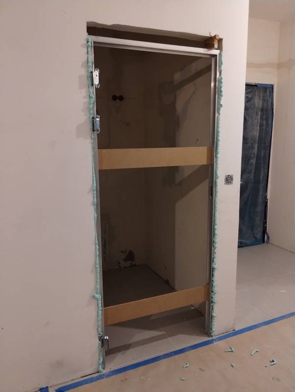 Ościeżnica drzwiowa zamontowana w surowym otworze, uszczelniona pianką montażową, zabezpieczona drewnianymi rozpórkami. Widoczne ślady prac remontowych na ścianach i podłodze.