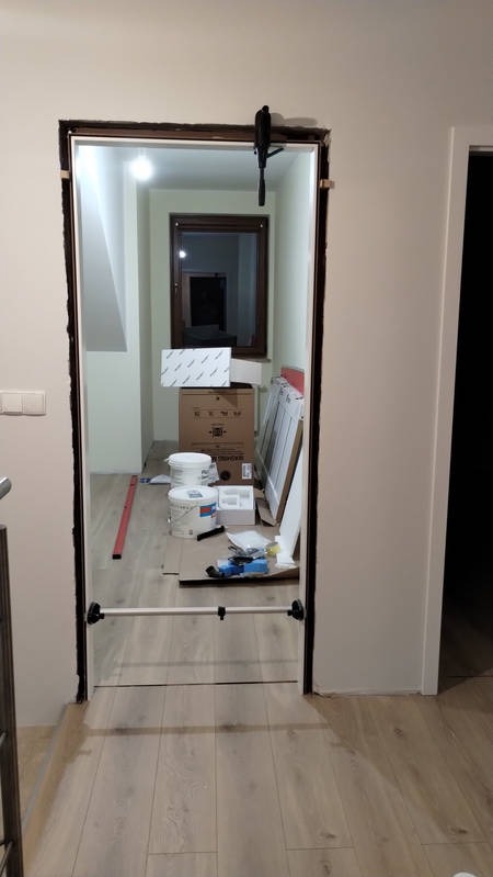 Przygotowanie do osadzenia futryny drzwiowej: widoczne narzędzia montażowe, poziomica oraz wstępnie przygotowane ościeże z podkładkami stabilizującymi, w tle widoczne materiały budowlane.