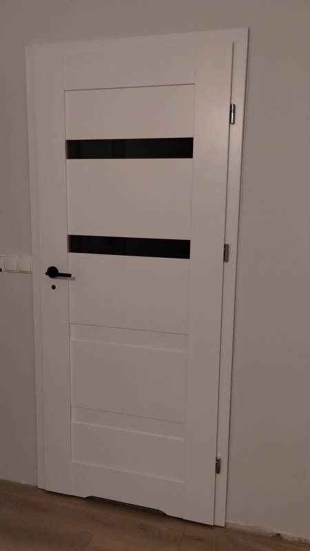 Białe drzwi wewnętrzne z czarnymi, poziomymi przeszkleniami, zamontowane w szarej ścianie, widoczna czarna klamka i fragment drewnianej podłogi.
