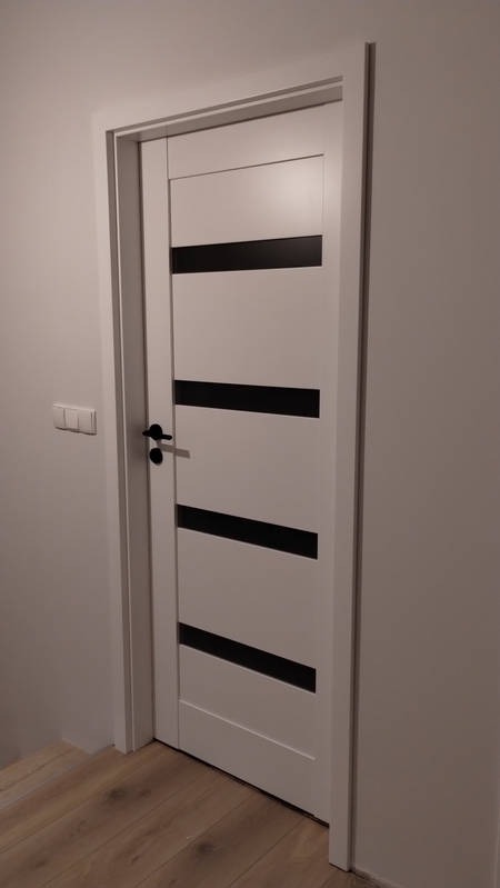 Białe drzwi wewnętrzne z czarnymi, poziomymi wstawkami, zamontowane w białej ościeżnicy, widoczne z boku, na tle białej ściany i drewnianej podłogi.