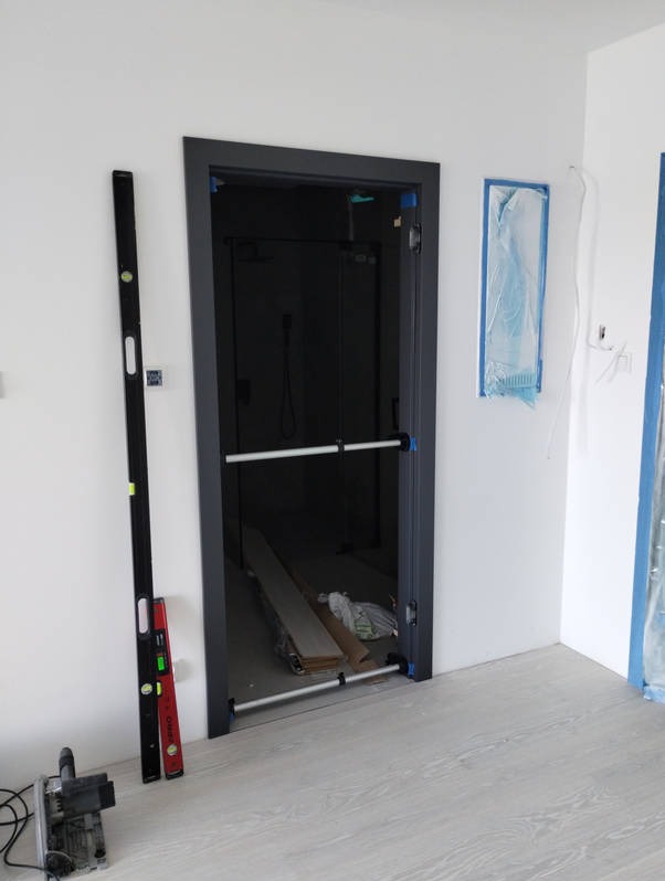 Ciemna ościeżnica z zamontowanymi rozpórkami, widoczna wnęka z materiałami budowlanymi, poziomice oparte o ścianę, piła ukosowa na podłodze, zabezpieczone folią fragmenty ścian.