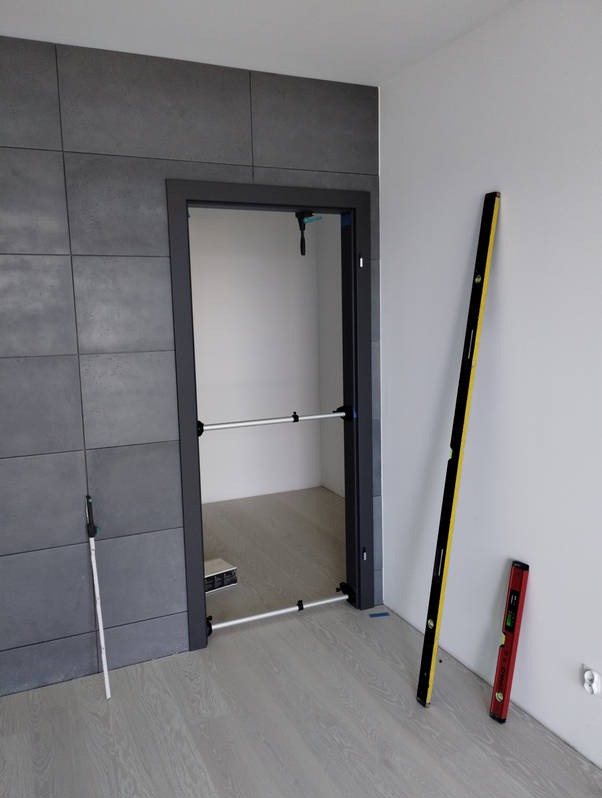 Szara ościeżnica drzwiowa z rozpórkami teleskopowymi podczas montażu, poziomica pionowa oparta o ścianę, płytki ceramiczne na ścianie po lewej stronie.