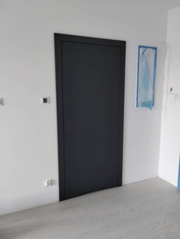 Nowoczesne, grafitowe drzwi wewnętrzne z minimalistyczną ościeżnicą, zamontowane w białej ścianie nad jasnymi panelami podłogowymi. Widoczna osłona zabezpieczająca na ścianie.