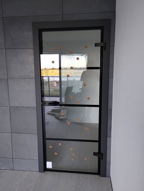 Szklane drzwi wewnętrzne z czarnymi szprosami, zabezpieczone folią ochronną, zamontowane w szarej ościeżnicy, widok na jasne panele podłogowe i szare płytki ścienne.