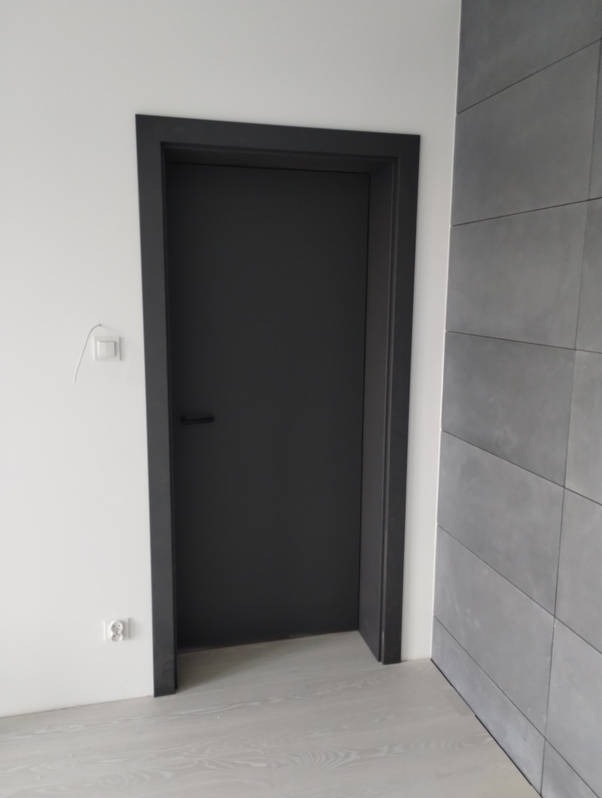 Zamknięte, nowoczesne, grafitowe drzwi z czarną ościeżnicą w minimalistycznym wnętrzu z białą ścianą i szarymi płytkami imitującymi beton.