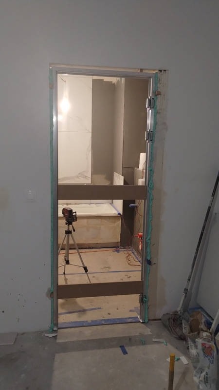 W trakcie montażu ościeżnicy drzwiowej w Gdańsku, widoczna piana montażowa uszczelniająca ramę, w tle wykończenie łazienki z wanną i laserowy niwelator na statywie.