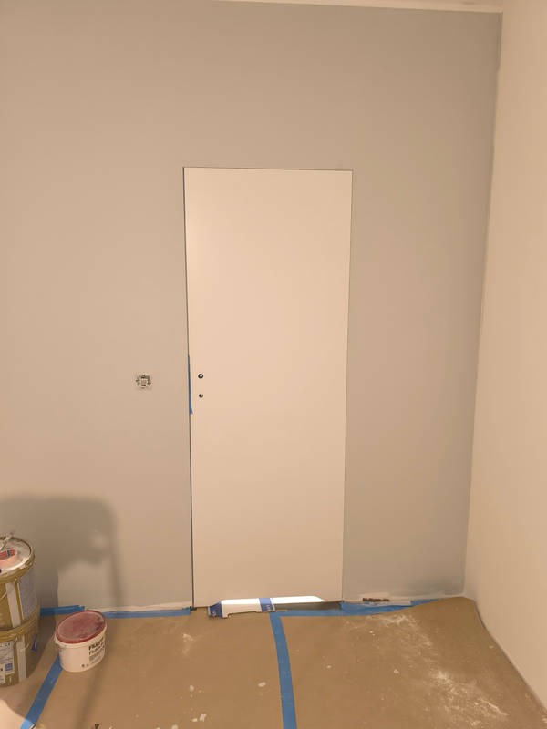 Białe, minimalistyczne drzwi wewnętrzne w trakcie montażu, zabezpieczone niebieską taśmą malarską, widoczne puszki z farbą i zabezpieczona podłoga.