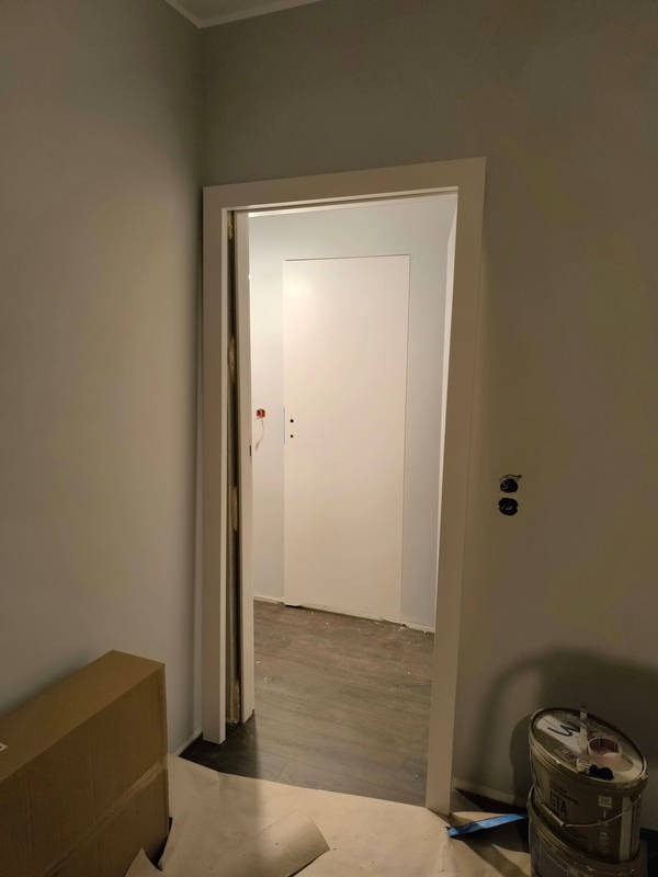 Białe, minimalistyczne drzwi wewnętrzne z ościeżnicą, widoczne z perspektywy wnętrza, z kartonem po lewej i puszkami z farbą po prawej stronie, na podłodze panele.