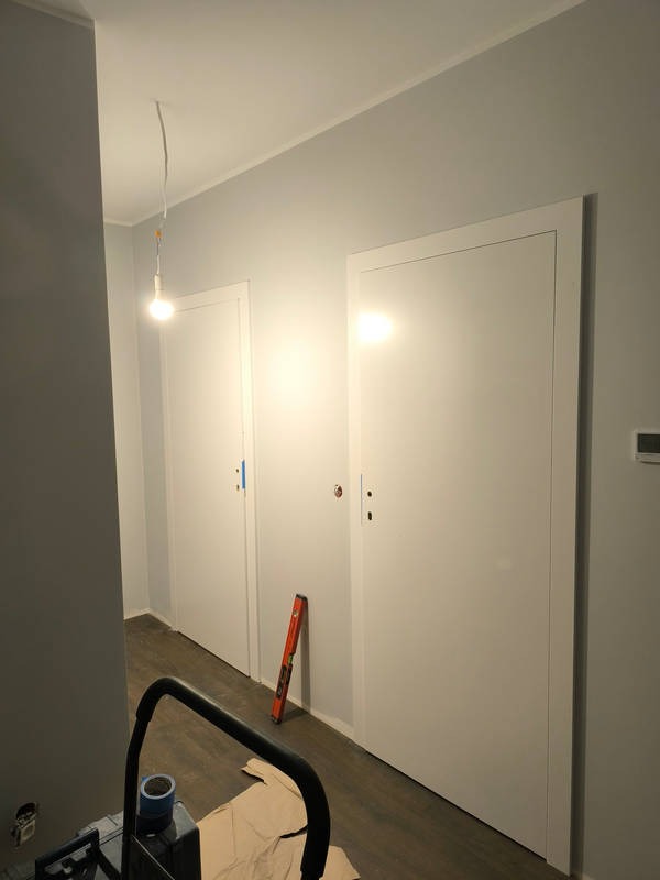 Dwa białe, minimalistyczne skrzydła drzwiowe zamontowane w szarej ścianie, poziomica oparta o framugę, widok z góry na rozłożony karton ochronny na podłodze i fragment drabiny malarskiej