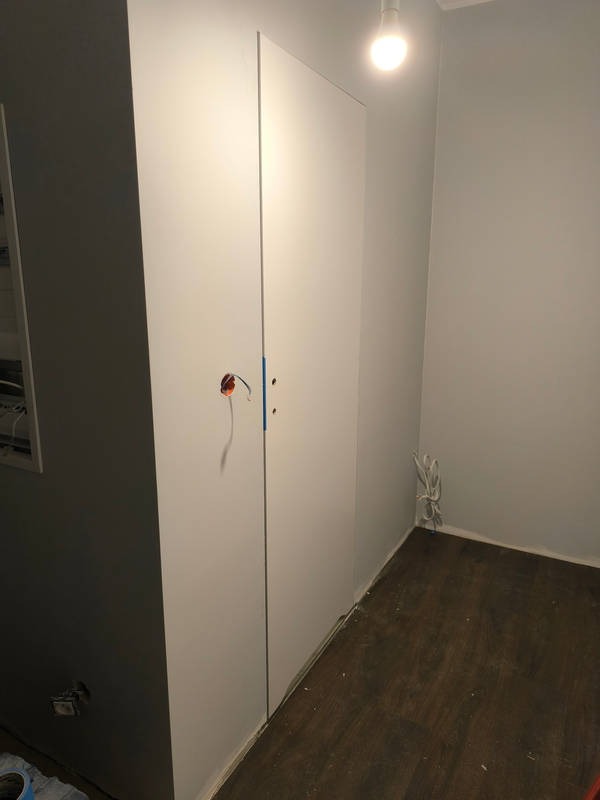 Biała szafa wnękowa z widocznym otworem na gniazdko elektryczne i wystającymi przewodami, zamontowana na podłodze z paneli imitujących ciemne drewno.