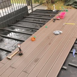 Bax-bud - Montaż deski kompozytowej na tarasie zewnętrznym. Widoczne narzędzia: młotek, wkrętarka. Częściowo ułożona podłoga z desek w kolorze brązowym. W tle metalowe ogrodzenie.