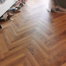 Bax-bud - Ułożona podłoga z paneli w jodełkę, widoczne resztki materiałów i zabezpieczone meble folią, jasne wnętrze z widokiem na balkon.