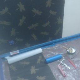 Bax-bud - Tapeta w odcieniach szarości z motywem ważek, wałek do tapetowania, pędzel i wiadro z klejem na folii ochronnej. Widoczny fragment ściany z tapetą.