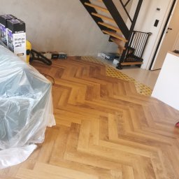 Bax-bud - Parkiet ułożony w jodełkę, widok na schody z drewnianymi stopniami i czarną metalową konstrukcją. Częściowo zakryty folią ochronną, w tle ściana i elementy wykończeniowe.