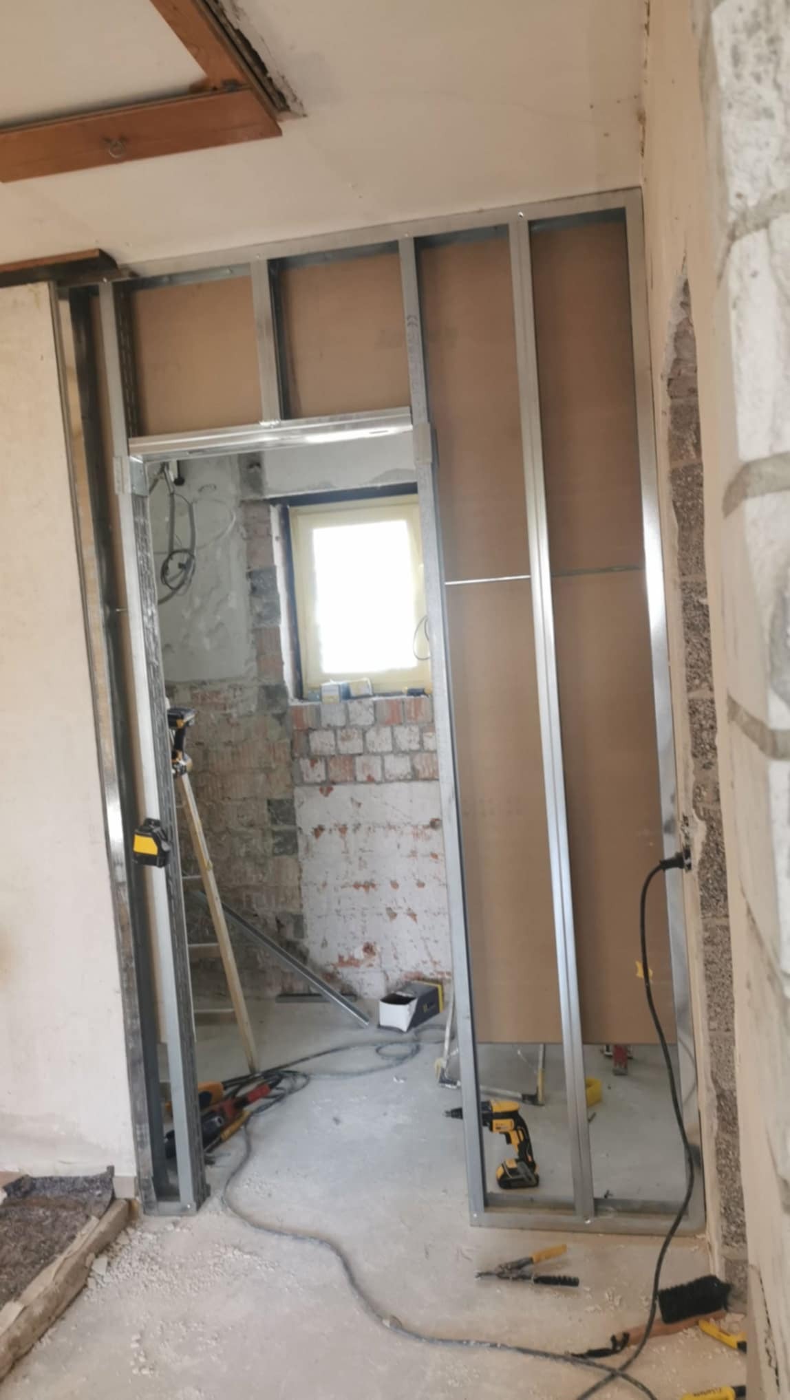 Montaż metalowej konstrukcji pod płytę G-K w remontowanym wnętrzu z widoczną starą cegłą. W tle okno z jasnym światłem, narzędzia na podłodze, drabina.