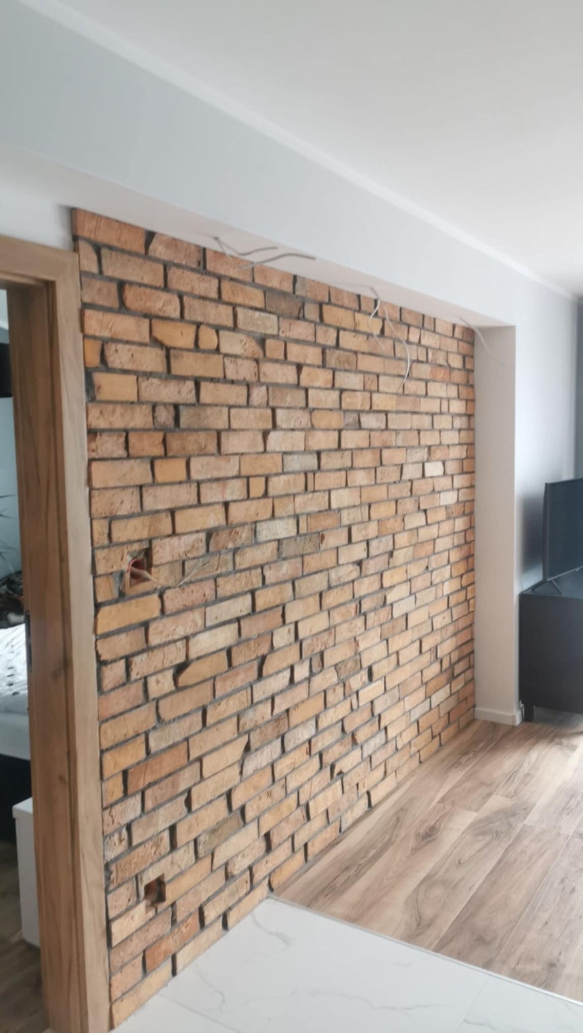 Dekoracyjna ściana z cegły klinkierowej w salonie, widoczne przewody elektryczne, drewniana podłoga i fragment mebli. Nietypowe wykończenie wnętrza z akcentem rustykalnym.