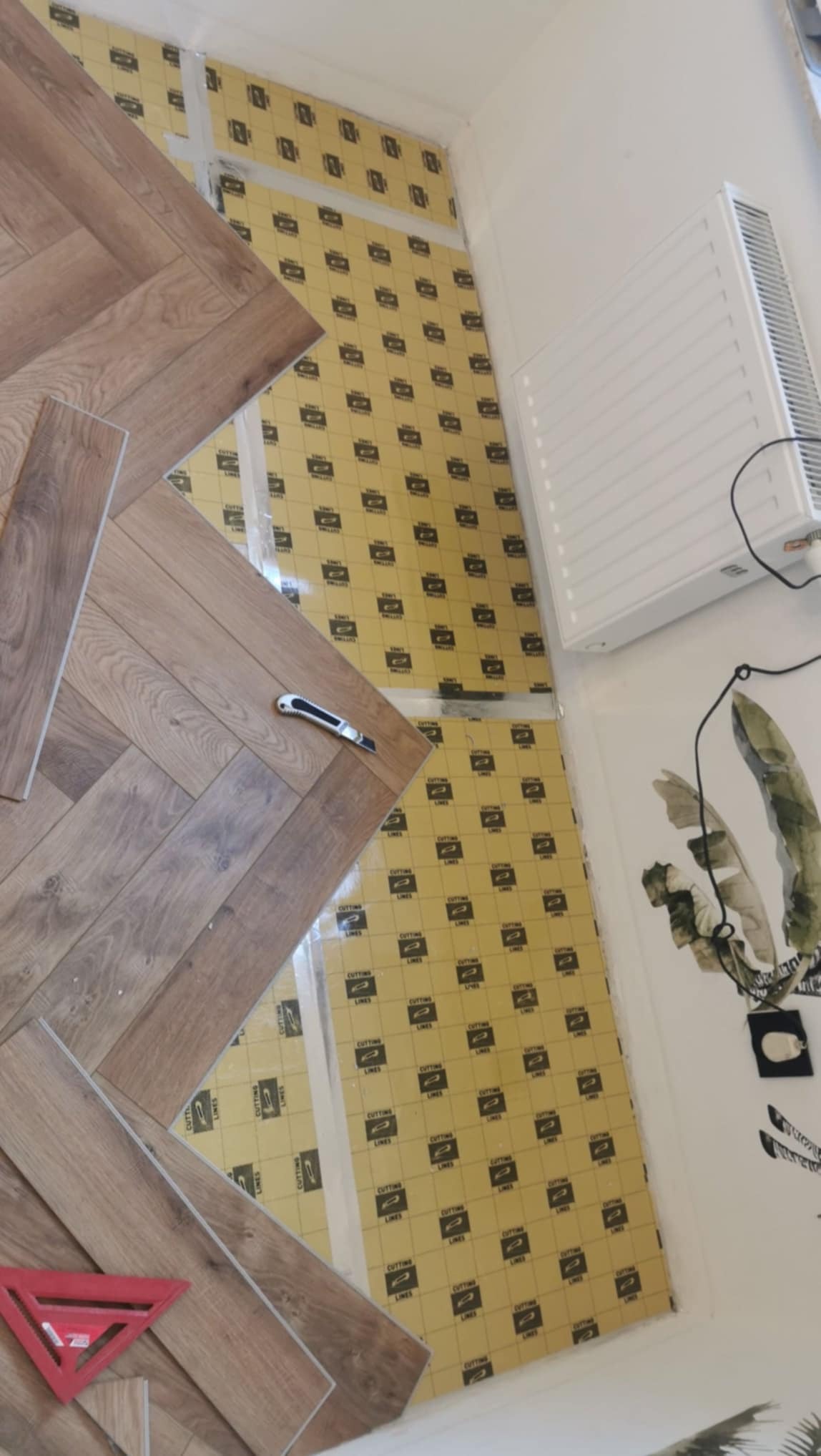 Układanie paneli podłogowych w jodełkę, widoczne narzędzie do cięcia i fragment podkładu izolacyjnego z nadrukiem. Częściowo ułożona podłoga w jasnym wnętrzu.