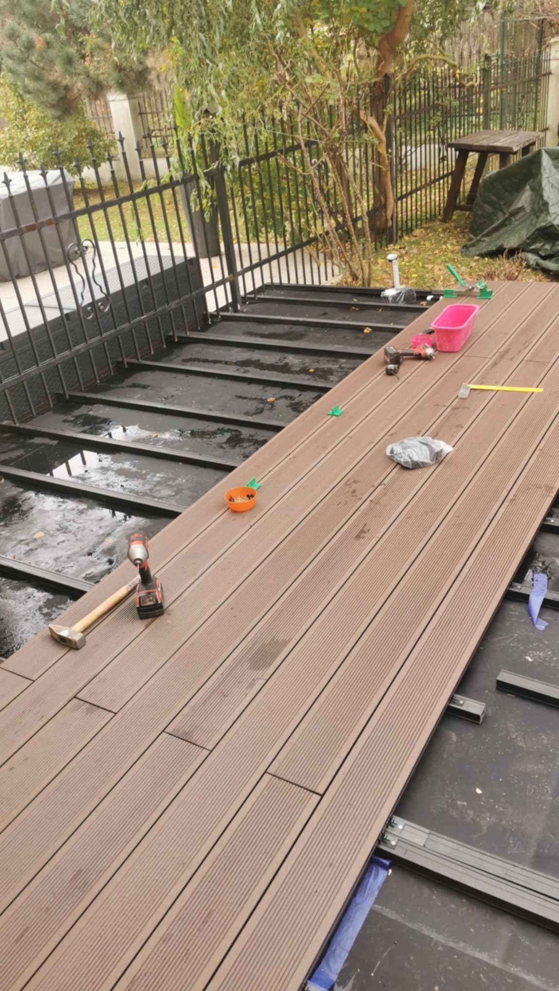 Montaż deski kompozytowej na tarasie zewnętrznym. Widoczne narzędzia: młotek, wkrętarka. Częściowo ułożona podłoga z desek w kolorze brązowym. W tle metalowe ogrodzenie.