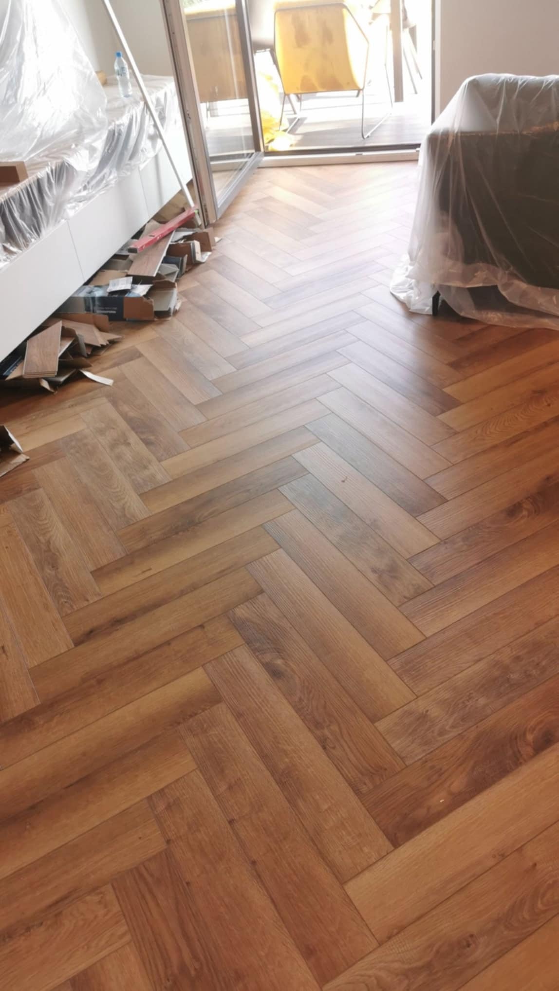 Ułożona podłoga z paneli w jodełkę, widoczne resztki materiałów i zabezpieczone meble folią, jasne wnętrze z widokiem na balkon.