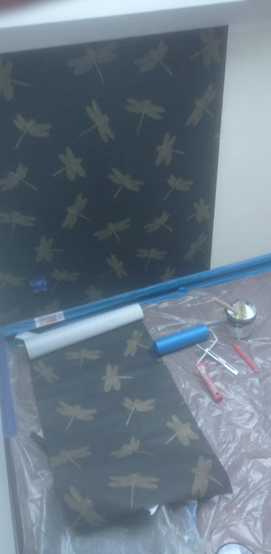 Tapeta w odcieniach szarości z motywem ważek, wałek do tapetowania, pędzel i wiadro z klejem na folii ochronnej. Widoczny fragment ściany z tapetą.