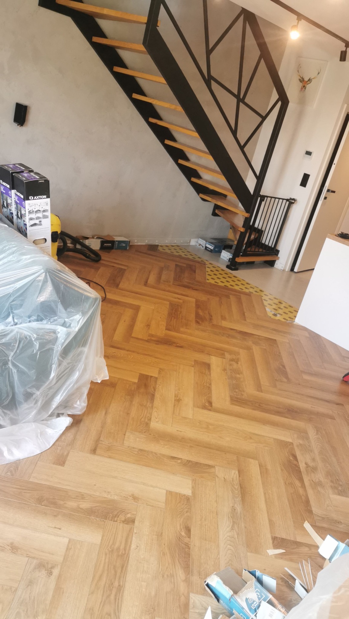 Parkiet ułożony w jodełkę, widok na schody z drewnianymi stopniami i czarną metalową konstrukcją. Częściowo zakryty folią ochronną, w tle ściana i elementy wykończeniowe.