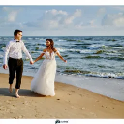 Para młoda, boso, idzie brzegiem morza trzymając się za ręce. Panna młoda w białej sukni, pan młody w białej koszuli i czarnych spodniach. Słońce, fale, piasek.