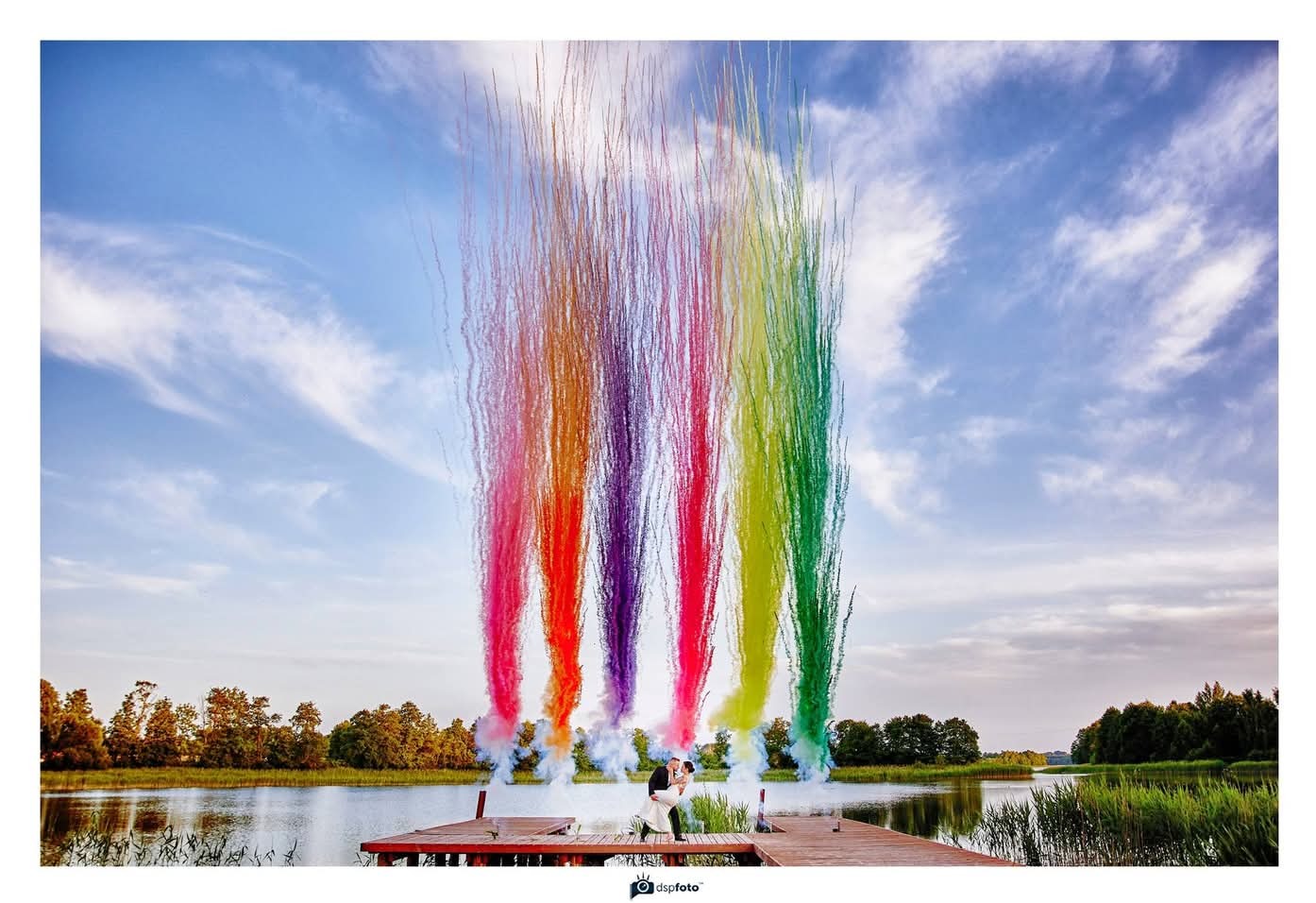 Para młoda w Suwałkach na drewnianym pomoście nad jeziorem, obejmująca się pod tęczowym deszczem kolorowych fontann pirotechnicznych, w tle błękitne niebo z chmurami.