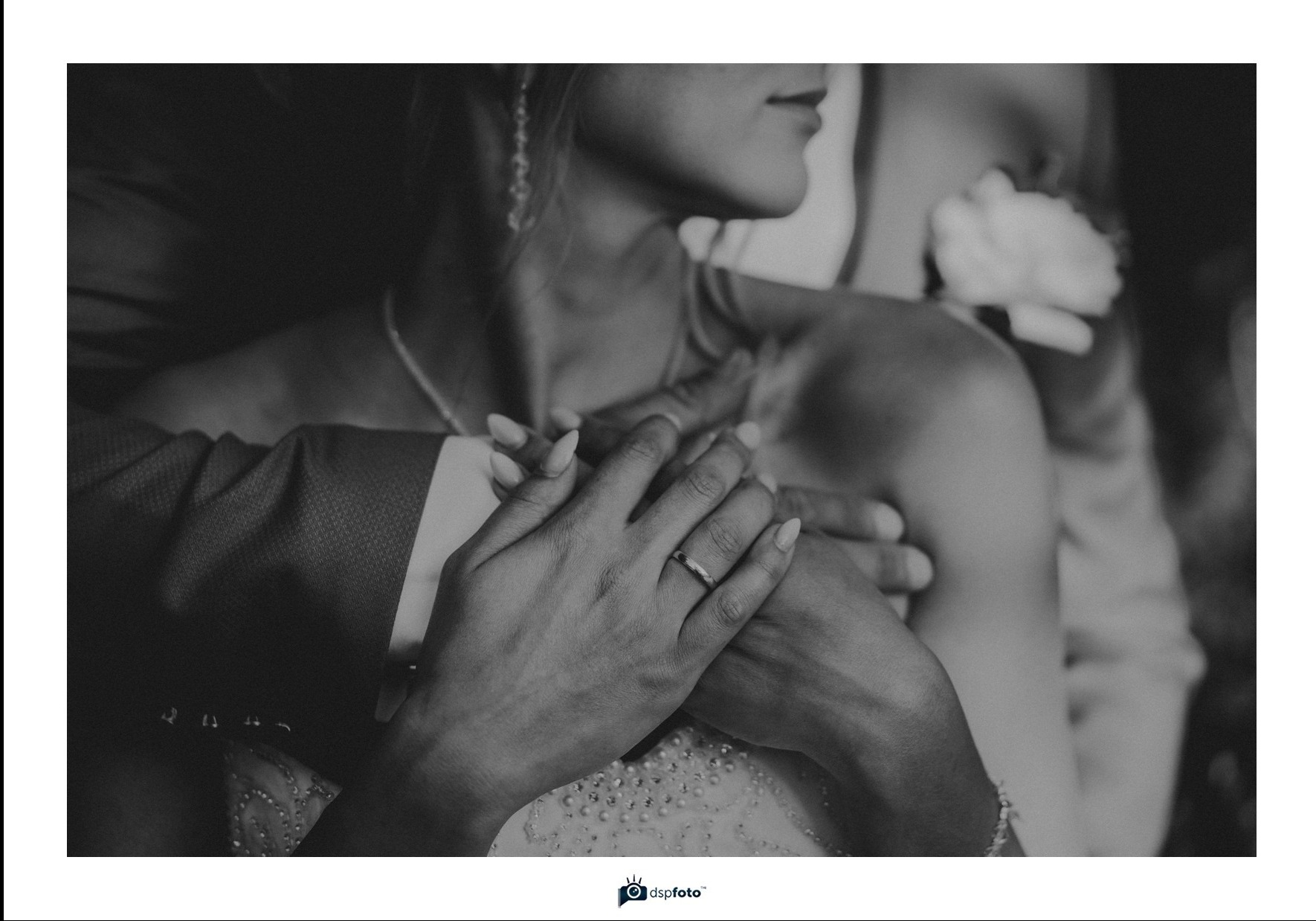 Czarno-białe zbliżenie dłoni pary młodej z pierścionkiem na palcu kobiety, delikatnie splecione dłonie na tle sukni ślubnej i marynarki pana młodego.